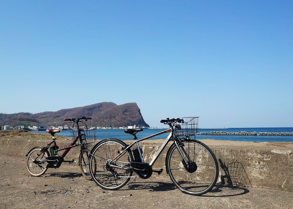 青い空と海、シリパ岬を背景に止まる2台の電動クロスバイクの写真